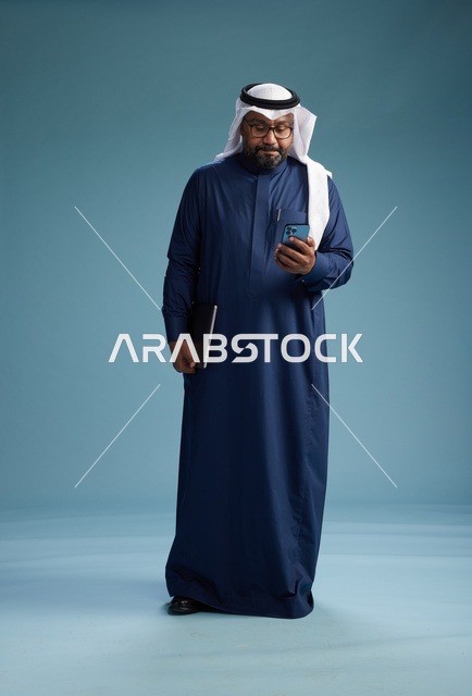 بورتريه لرجل سعودي مبتسم  يرتدي ثوب كحلي وغتره بيضاء يحمل لابتوب وهاتف ذكي, استخدام التقنيه الحديثه, كامل الجسم, خلفيه زرقاء