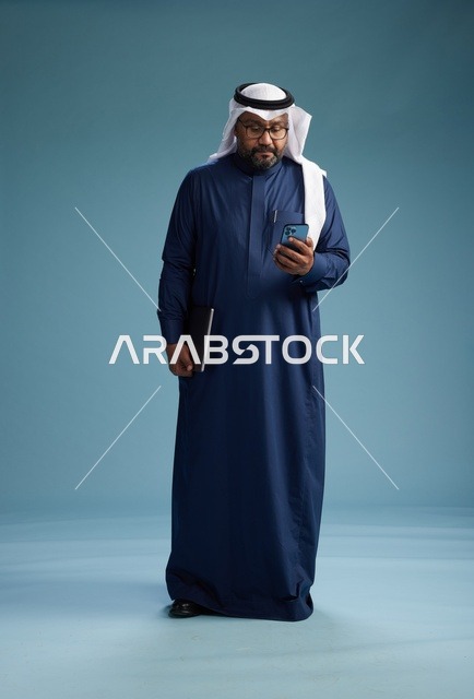 بورتريه لرجل سعودي مبتسم  يرتدي ثوب كحلي وغتره بيضاء يحمل لابتوب وهاتف ذكي, استخدام التقنيه الحديثه, كامل الجسم, خلفيه زرقاء