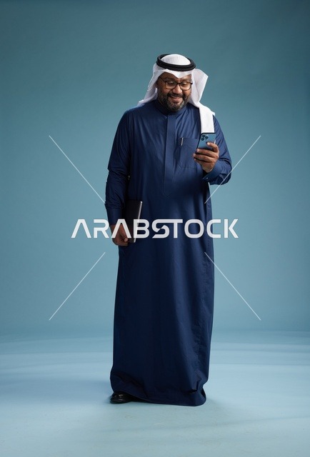 بورتريه لرجل سعودي مبتسم  يرتدي ثوب كحلي وغتره بيضاء يحمل لابتوب وهاتف ذكي, استخدام التقنيه الحديثه, كامل الجسم, خلفيه زرقاء