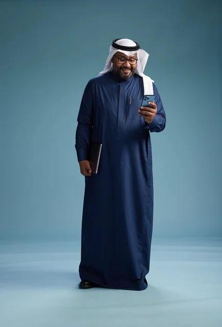 A portrait of a smiling Saudi man wearing a navy blue thobe and a white ghutrah, holding a laptop and a smartphone, using modern technology, full body, with a blue background.
