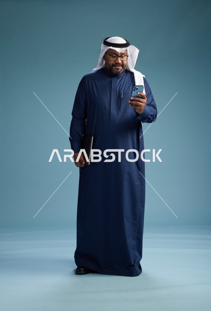 بورتريه لرجل سعودي مبتسم  يرتدي ثوب كحلي وغتره بيضاء يحمل لابتوب وهاتف ذكي, استخدام التقنيه الحديثه, كامل الجسم, خلفيه زرقاء