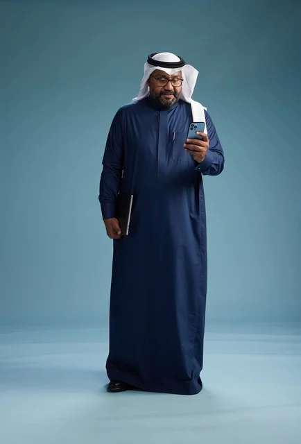 A portrait of a smiling Saudi man wearing a navy blue thobe and a white ghutrah, holding a laptop and a smartphone, using modern technology, full body, with a blue background.