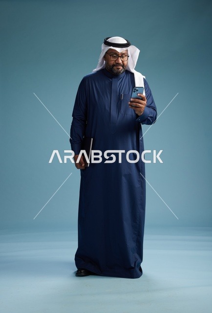 بورتريه لرجل سعودي مبتسم  يرتدي ثوب كحلي وغتره بيضاء يحمل لابتوب وهاتف ذكي, استخدام التقنيه الحديثه, كامل الجسم, خلفيه زرقاء