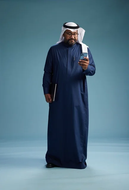 A portrait of a smiling Saudi man wearing a navy blue thobe and a white ghutrah, holding a laptop and a smartphone, using modern technology, full body, with a blue background. A portrait of a smiling Saudi man wearing a navy blue thobe and a white ghutrah, holding a laptop and a smartphone, using modern technology, full body, with a blue background.