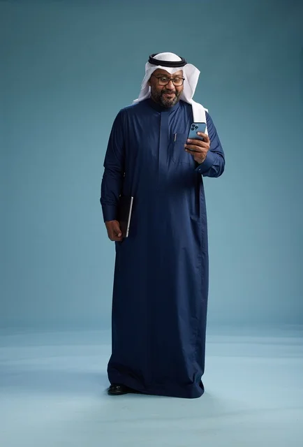 A portrait of a smiling Saudi man wearing a navy blue thobe and a white ghutrah, holding a laptop and a smartphone, using modern technology, full body, with a blue background.