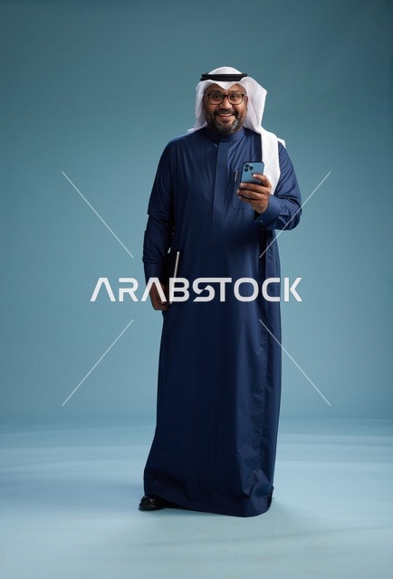 بورتريه لرجل سعودي مبتسم  يرتدي ثوب كحلي وغتره بيضاء يحمل لابتوب وهاتف ذكي, استخدام التقنيه الحديثه, كامل الجسم, خلفيه زرقاء
