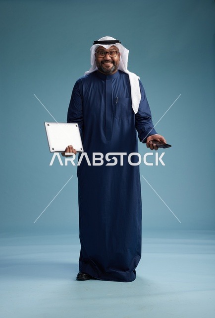 بورتريه لرجل سعودي مبتسم  يرتدي ثوب كحلي وغتره بيضاء يحمل لابتوب وهاتف ذكي, استخدام التقنيه الحديثه, كامل الجسم, خلفيه زرقاء