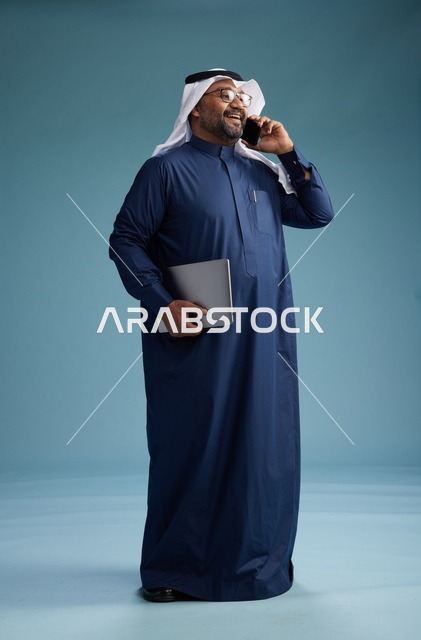 بورتريه لرجل عربي سعودي خليجي يرتدي ثوب كحلي وغتره بيضاء يستخدم الهاتف المحمول ويمسك جهاز التبليت باليد الاخرى, اجراء مكالمه هاتفيه, الجسم بالطول الكامل, خلفيه زرقاء