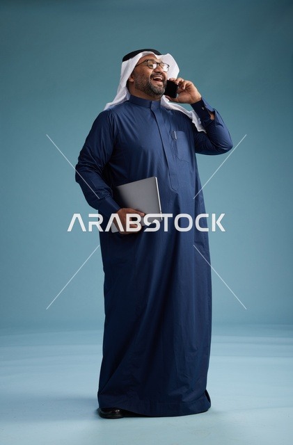 بورتريه لرجل عربي سعودي خليجي يرتدي ثوب كحلي وغتره بيضاء يستخدم الهاتف المحمول ويمسك جهاز التبليت باليد الاخرى, اجراء مكالمه هاتفيه, الجسم بالطول الكامل, خلفيه زرقاء