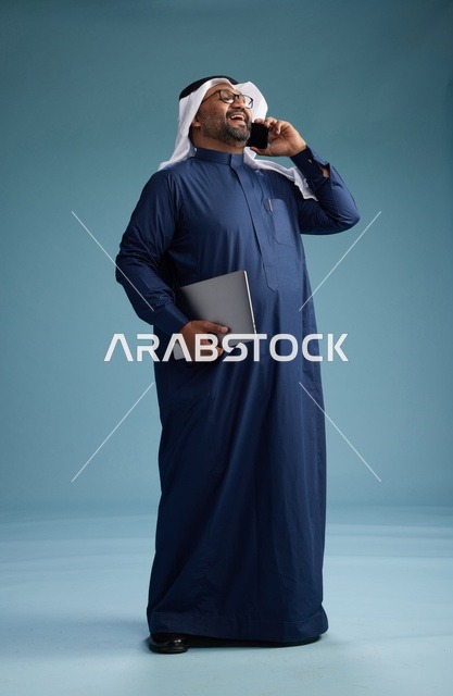 بورتريه لرجل عربي سعودي خليجي يرتدي ثوب كحلي وغتره بيضاء يستخدم الهاتف المحمول ويمسك جهاز التبليت باليد الاخرى, اجراء مكالمه هاتفيه, الجسم بالطول الكامل, خلفيه زرقاء