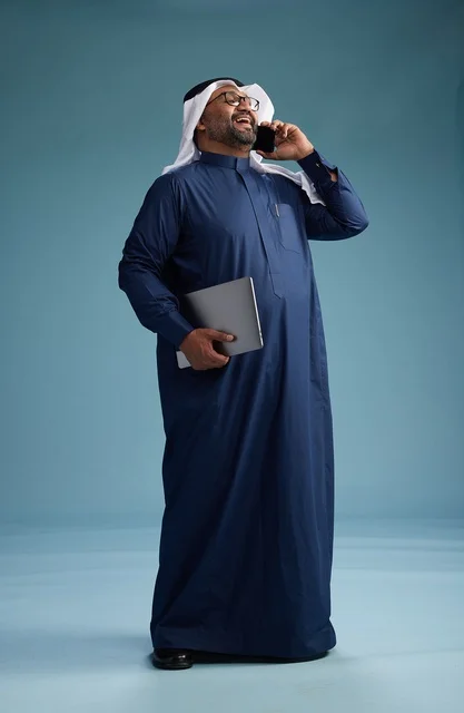 A portrait of an Arab Saudi Gulf man wearing a navy blue thobe and a white ghutrah, using a mobile phone and holding a tablet in the other hand, making a phone call, full body, blue background.