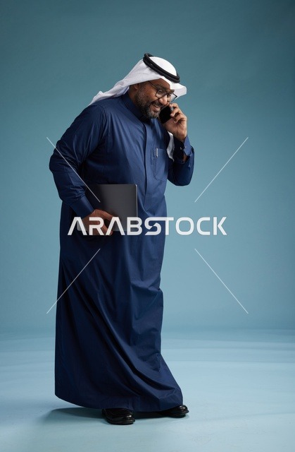 بورتريه لرجل عربي سعودي خليجي يرتدي ثوب كحلي وغتره بيضاء يستخدم الهاتف المحمول ويمسك جهاز التبليت باليد الاخرى, اجراء مكالمه هاتفيه, الجسم بالطول الكامل, خلفيه زرقاء