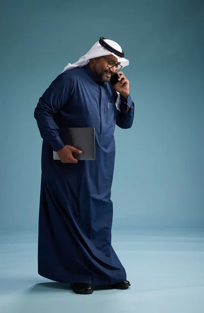 A portrait of an Arab Saudi Gulf man wearing a navy blue thobe and a white ghutrah, using a mobile phone and holding a tablet in the other hand, making a phone call, full body, blue background.
