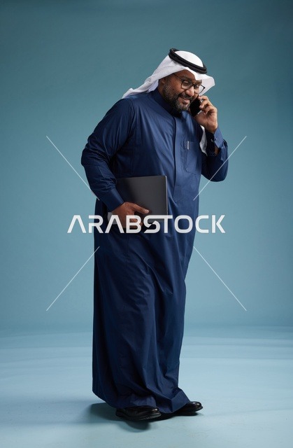 بورتريه لرجل عربي سعودي خليجي يرتدي ثوب كحلي وغتره بيضاء يستخدم الهاتف المحمول ويمسك جهاز التبليت باليد الاخرى, اجراء مكالمه هاتفيه, الجسم بالطول الكامل, خلفيه زرقاء