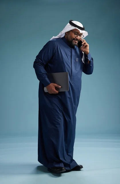 بورتريه لرجل عربي سعودي خليجي يرتدي ثوب كحلي وغتره بيضاء يستخدم الهاتف المحمول ويمسك جهاز التبليت باليد الاخرى, اجراء مكالمه هاتفيه, الجسم بالطول الكامل, خلفيه زرقاء