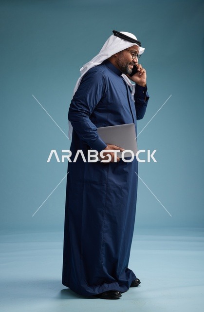 بورتريه لرجل عربي سعودي خليجي يرتدي ثوب كحلي وغتره بيضاء يستخدم الهاتف المحمول ويمسك جهاز التبليت باليد الاخرى, اجراء مكالمه هاتفيه, الجسم بالطول الكامل, خلفيه زرقاء