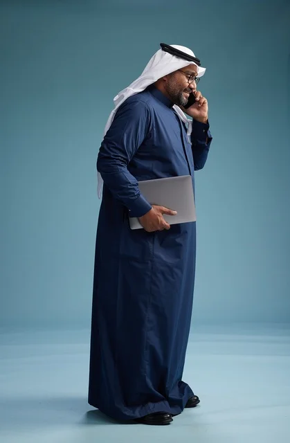 بورتريه لرجل عربي سعودي خليجي يرتدي ثوب كحلي وغتره بيضاء يستخدم الهاتف المحمول ويمسك جهاز التبليت باليد الاخرى, اجراء مكالمه هاتفيه, الجسم بالطول الكامل, خلفيه زرقاء