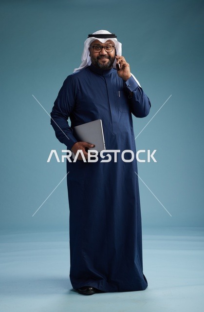 بورتريه لرجل عربي سعودي خليجي يرتدي ثوب كحلي وغتره بيضاء يستخدم الهاتف المحمول ويمسك جهاز التبليت باليد الاخرى, اجراء مكالمه هاتفيه, الجسم بالطول الكامل, خلفيه زرقاء