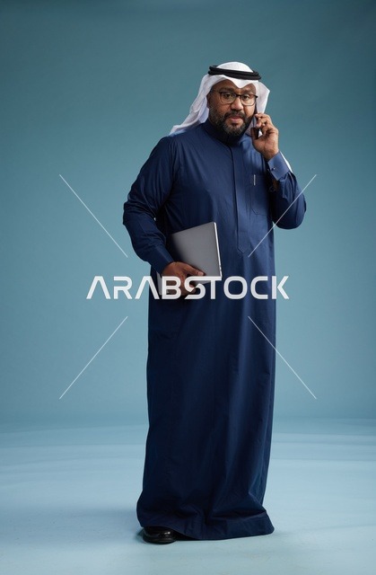 بورتريه لرجل عربي سعودي خليجي يرتدي ثوب كحلي وغتره بيضاء يستخدم الهاتف المحمول ويمسك جهاز التبليت باليد الاخرى, اجراء مكالمه هاتفيه, الجسم بالطول الكامل, خلفيه زرقاء