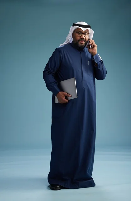 بورتريه لرجل عربي سعودي خليجي يرتدي ثوب كحلي وغتره بيضاء يستخدم الهاتف المحمول ويمسك جهاز التبليت باليد الاخرى, اجراء مكالمه هاتفيه, الجسم بالطول الكامل, خلفيه زرقاء