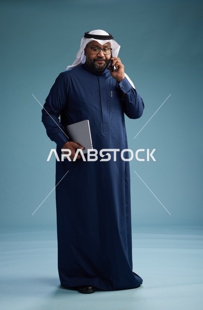 بورتريه لرجل عربي سعودي خليجي يرتدي ثوب كحلي وغتره بيضاء يستخدم الهاتف المحمول ويمسك جهاز التبليت باليد الاخرى, اجراء مكالمه هاتفيه, الجسم بالطول الكامل, خلفيه زرقاء