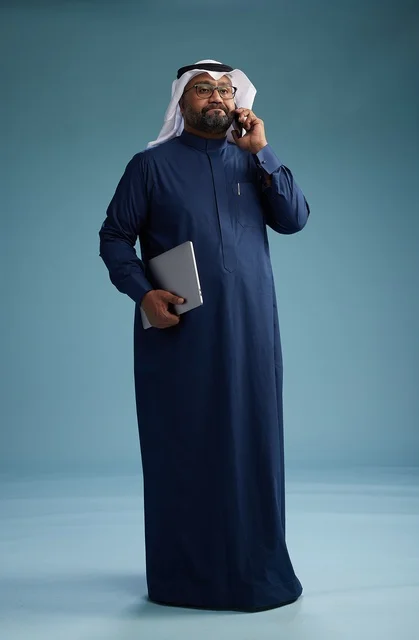 A portrait of an Arab Saudi Gulf man wearing a navy blue thobe and a white ghutrah, using a mobile phone and holding a tablet in the other hand, making a phone call, full body, blue background.