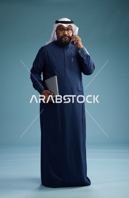 بورتريه لرجل عربي سعودي خليجي يرتدي ثوب كحلي وغتره بيضاء يستخدم الهاتف المحمول ويمسك جهاز التبليت باليد الاخرى, اجراء مكالمه هاتفيه, الجسم بالطول الكامل, خلفيه زرقاء