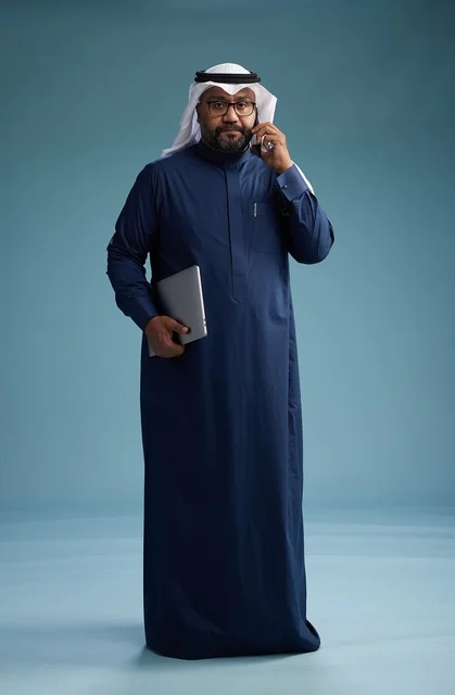 A portrait of an Arab Saudi Gulf man wearing a navy blue thobe and a white ghutrah, using a mobile phone and holding a tablet in the other hand, making a phone call, full body, blue background.