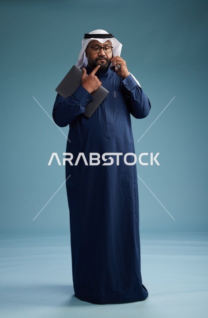 بورتريه لرجل عربي سعودي خليجي يرتدي ثوب كحلي وغتره بيضاء يستخدم الهاتف المحمول ويمسك جهاز التبليت باليد الاخرى, اجراء مكالمه هاتفيه, الجسم بالطول الكامل, خلفيه زرقاء