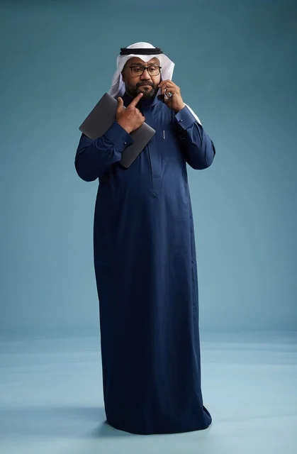A portrait of an Arab Saudi Gulf man wearing a navy blue thobe and a white ghutrah, using a mobile phone and holding a tablet in the other hand, making a phone call, full body, blue background.