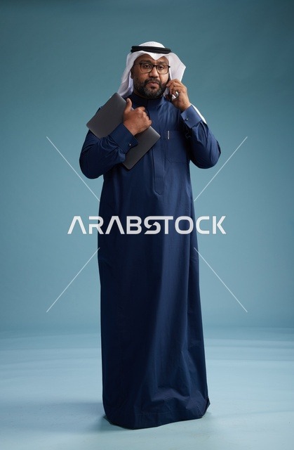 بورتريه لرجل عربي سعودي خليجي يرتدي ثوب كحلي وغتره بيضاء يستخدم الهاتف المحمول ويمسك جهاز التبليت باليد الاخرى, اجراء مكالمه هاتفيه, الجسم بالطول الكامل, خلفيه زرقاء