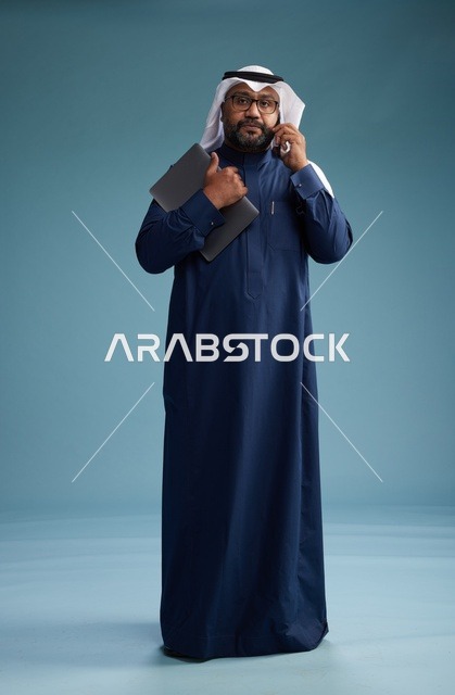 بورتريه لرجل عربي سعودي خليجي يرتدي ثوب كحلي وغتره بيضاء يستخدم الهاتف المحمول ويمسك جهاز التبليت باليد الاخرى, اجراء مكالمه هاتفيه, الجسم بالطول الكامل, خلفيه زرقاء