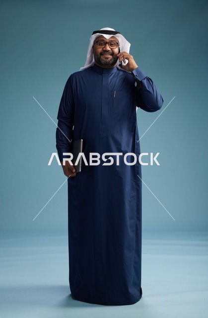 بورتريه لرجل عربي سعودي خليجي يرتدي ثوب كحلي وغتره بيضاء يستخدم الهاتف المحمول ويمسك جهاز التبليت باليد الاخرى, اجراء مكالمه هاتفيه, الجسم بالطول الكامل, خلفيه زرقاء