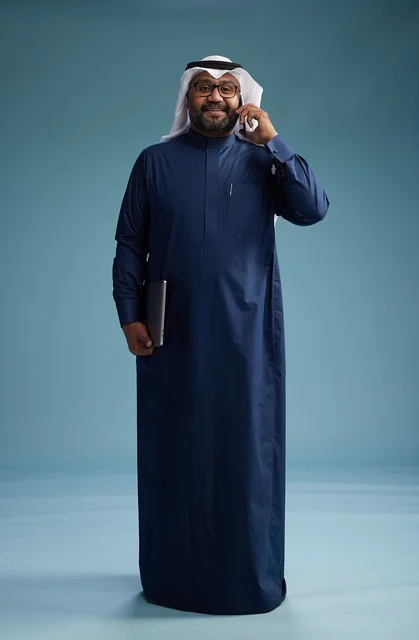 A portrait of an Arab Saudi Gulf man wearing a navy blue thobe and a white ghutrah, using a mobile phone and holding a tablet in the other hand, making a phone call, full body, blue background.