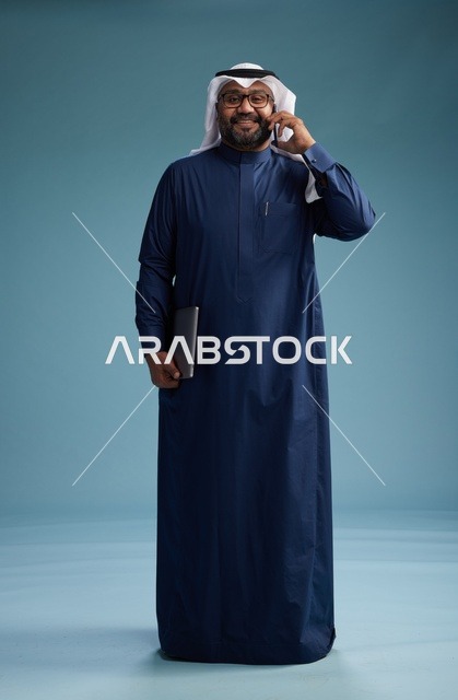 بورتريه لرجل عربي سعودي خليجي يرتدي ثوب كحلي وغتره بيضاء يستخدم الهاتف المحمول ويمسك جهاز التبليت باليد الاخرى, اجراء مكالمه هاتفيه, الجسم بالطول الكامل, خلفيه زرقاء