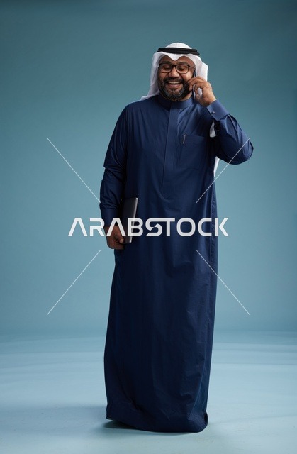 بورتريه لرجل عربي سعودي خليجي يرتدي ثوب كحلي وغتره بيضاء يستخدم الهاتف المحمول ويمسك جهاز التبليت باليد الاخرى, اجراء مكالمه هاتفيه, الجسم بالطول الكامل, خلفيه زرقاء