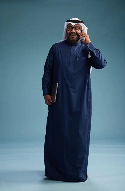 A portrait of an Arab Saudi Gulf man wearing a navy blue thobe and a white ghutrah, using a mobile phone and holding a tablet in the other hand, making a phone call, full body, blue background.