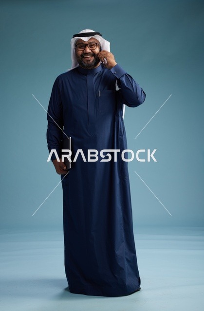 بورتريه لرجل عربي سعودي خليجي يرتدي ثوب كحلي وغتره بيضاء يستخدم الهاتف المحمول ويمسك جهاز التبليت باليد الاخرى, اجراء مكالمه هاتفيه, الجسم بالطول الكامل, خلفيه زرقاء