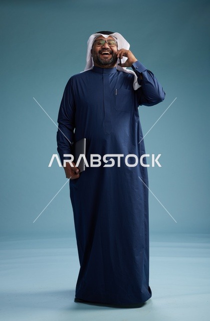 بورتريه لرجل عربي سعودي خليجي يرتدي ثوب كحلي وغتره بيضاء يستخدم الهاتف المحمول ويمسك جهاز التبليت باليد الاخرى, اجراء مكالمه هاتفيه, الجسم بالطول الكامل, خلفيه زرقاء