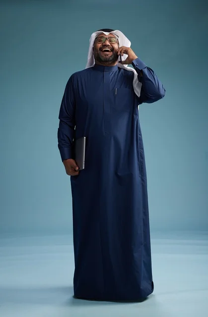 A portrait of an Arab Saudi Gulf man wearing a navy blue thobe and a white ghutrah, using a mobile phone and holding a tablet in the other hand, making a phone call, full body, blue background.