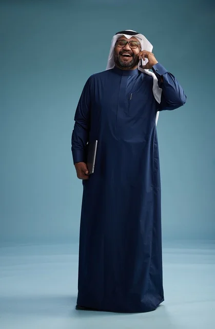 A portrait of an Arab Saudi Gulf man wearing a navy blue thobe and a white ghutrah, using a mobile phone and holding a tablet in the other hand, making a phone call, full body, blue background.
