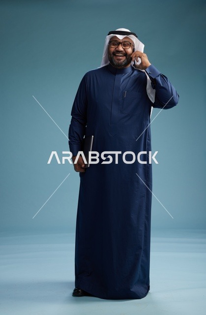 بورتريه لرجل عربي سعودي خليجي يرتدي ثوب كحلي وغتره بيضاء يستخدم الهاتف المحمول ويمسك جهاز التبليت باليد الاخرى, اجراء مكالمه هاتفيه, الجسم بالطول الكامل, خلفيه زرقاء