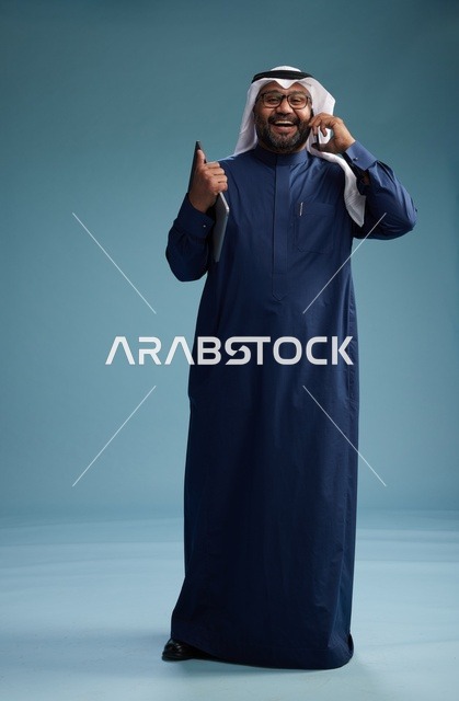 بورتريه لرجل عربي سعودي خليجي يرتدي ثوب كحلي وغتره بيضاء يستخدم الهاتف المحمول ويمسك جهاز التبليت باليد الاخرى, اجراء مكالمه هاتفيه, الجسم بالطول الكامل, خلفيه زرقاء