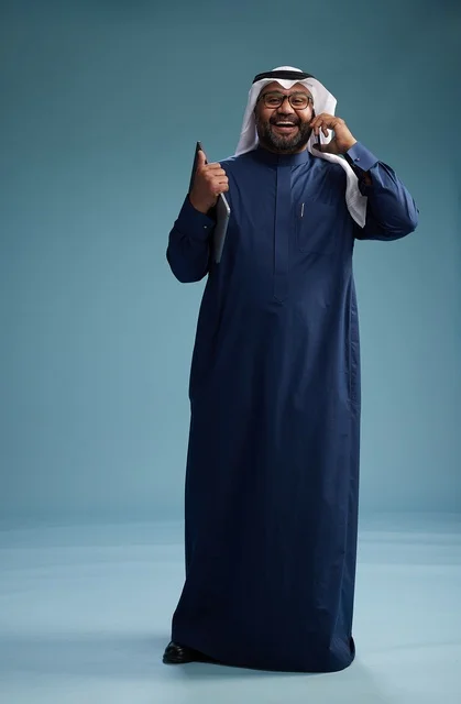 A portrait of an Arab Saudi Gulf man wearing a navy blue thobe and a white ghutrah, using a mobile phone and holding a tablet in the other hand, making a phone call, full body, blue background.