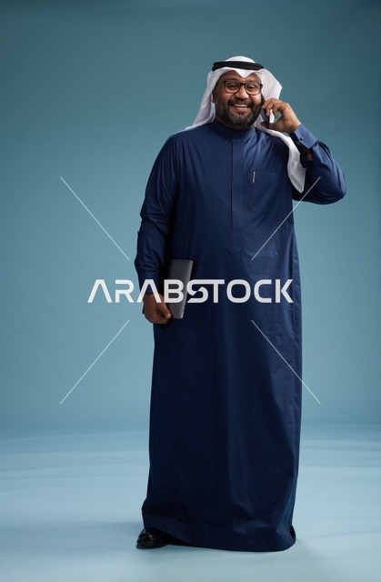 بورتريه لرجل عربي سعودي خليجي يرتدي ثوب كحلي وغتره بيضاء يستخدم الهاتف المحمول ويمسك جهاز التبليت باليد الاخرى, اجراء مكالمه هاتفيه, الجسم بالطول الكامل, خلفيه زرقاء
