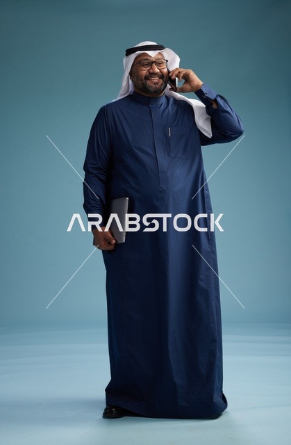 بورتريه لرجل عربي سعودي خليجي يرتدي ثوب كحلي وغتره بيضاء يستخدم الهاتف المحمول ويمسك جهاز التبليت باليد الاخرى, اجراء مكالمه هاتفيه, الجسم بالطول الكامل, خلفيه زرقاء