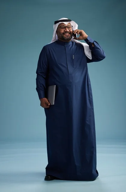 A portrait of an Arab Saudi Gulf man wearing a navy blue thobe and a white ghutrah, using a mobile phone and holding a tablet in the other hand, making a phone call, full body, blue background.