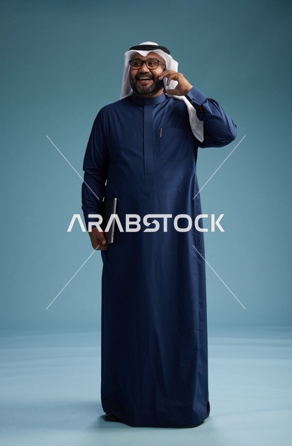 بورتريه لرجل عربي سعودي خليجي يرتدي ثوب كحلي وغتره بيضاء يستخدم الهاتف المحمول ويمسك جهاز التبليت باليد الاخرى, اجراء مكالمه هاتفيه, الجسم بالطول الكامل, خلفيه زرقاء