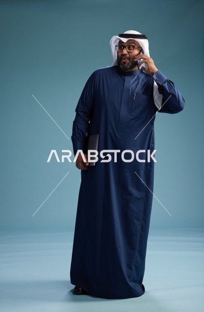 بورتريه لرجل عربي سعودي خليجي يرتدي ثوب كحلي وغتره بيضاء يستخدم الهاتف المحمول ويمسك جهاز التبليت باليد الاخرى, اجراء مكالمه هاتفيه, الجسم بالطول الكامل, خلفيه زرقاء