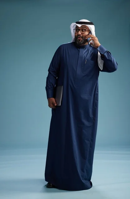 A portrait of an Arab Saudi Gulf man wearing a navy blue thobe and a white ghutrah, using a mobile phone and holding a tablet in the other hand, making a phone call, full body, blue background.